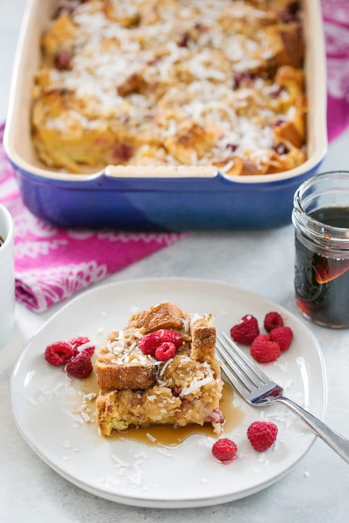 Overhead view of a plate with a serving of raspberry coconut french toast casserole with the baking dish in the background, along with a jar of maple syrup