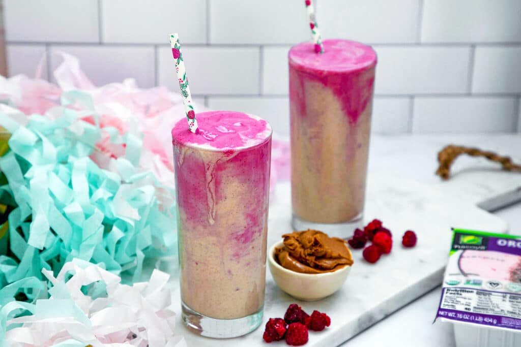 Landscape view of two almond butter and jelly smoothies with bowl of almond butter, frozen raspberries, and tofu package alongside