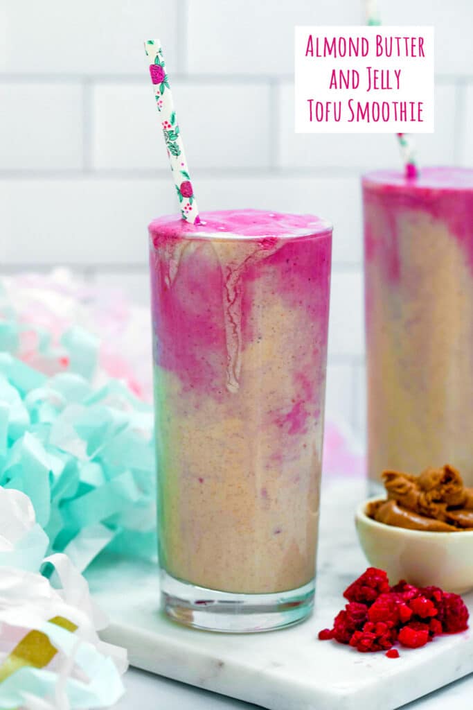 Head-on view of almond butter and tofu smoothie with frozen raspberries and almond butter alongside and second smoothie in the background and recipe title at top of image