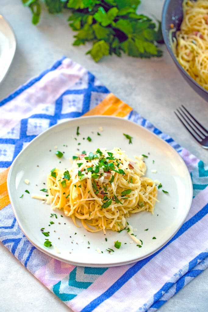 Overhead view of a grey plate of angel hair alfredo topped with parsley, pancetta, jalapeño, and parmesan with parsley in the background