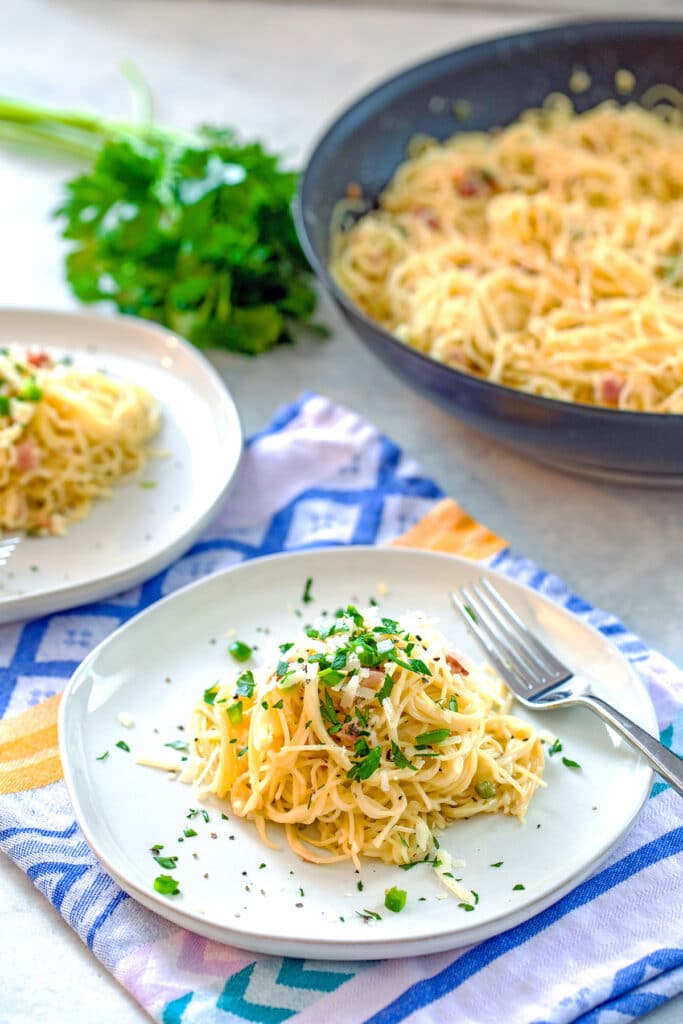 Plate of angel hair alfredo with jalapeño and pancetta with second plate, parsley, and skillet of pasta in background