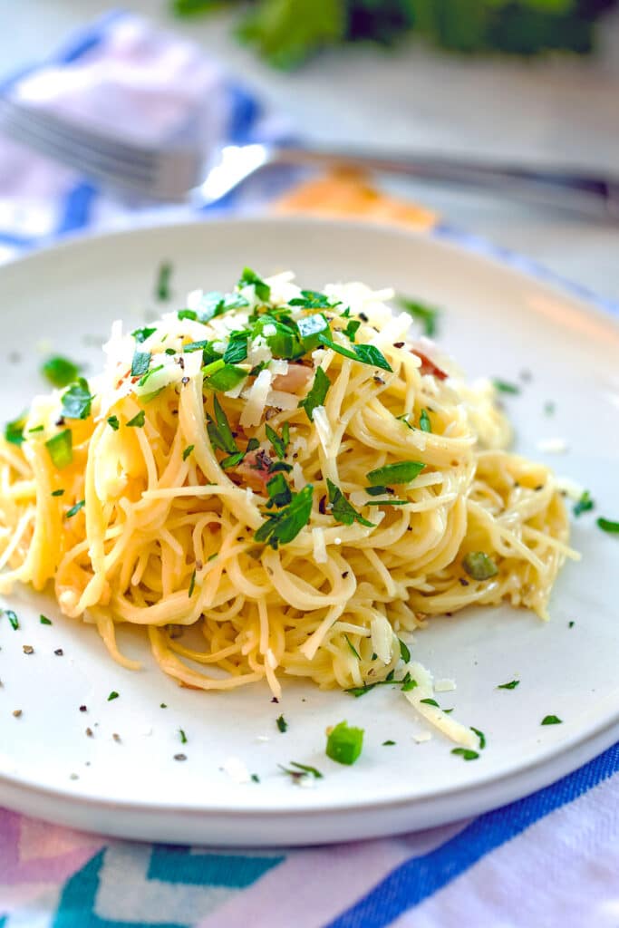 Closeup head-on image of angel hair alfredo on a plate topped with parmesan cheese, parsley, jalapeño, and pancetta