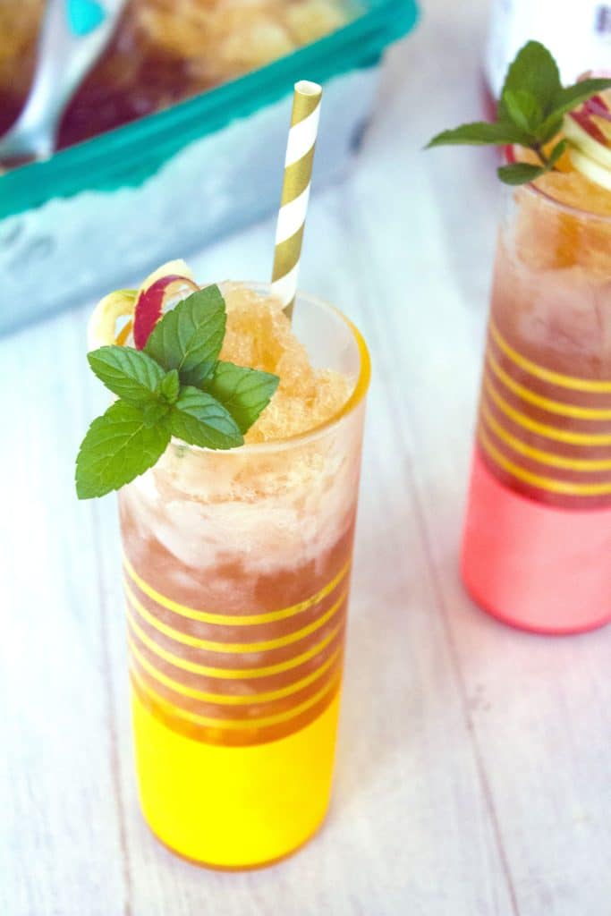 Overhead view of apple cider bourbon slush in a tall glass with mint and apple garnish and white and gold straw with second glass and container of bourbon slush in the background