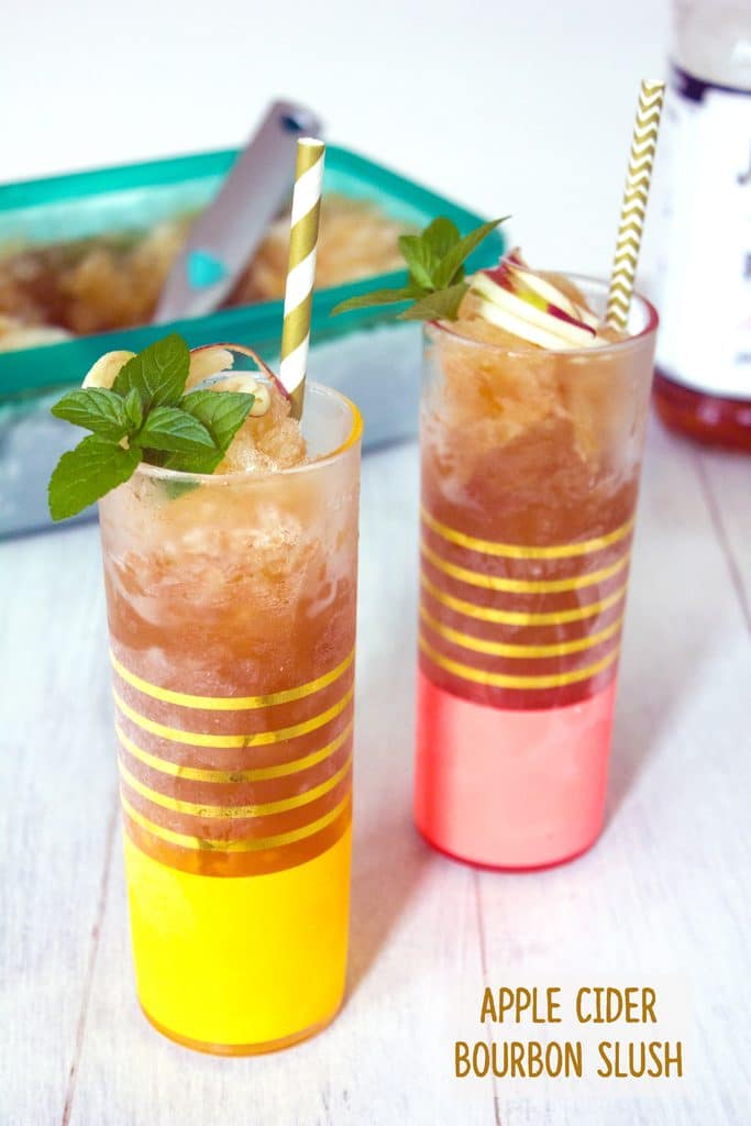 Head-on view of two tall glasses filled with apple cider bourbon slush with mint and apple garnish and gold and white straws on a white wood surface with container of bourbon slush in the background and recipe title at bottom