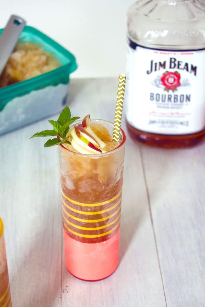 Head-on view of tall pink and gold class of apple cider bourbon slush with mint and apple garnish and white and gold straw with container of slush and bottle of Jim Beam Bourbon in background