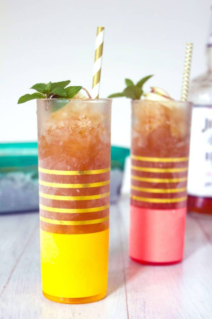 Head-on closeup view of yellow and gold glass of apple cider bourbon slush with mint garnish and white and gold straw with second glass in the background