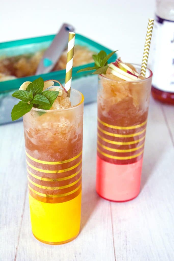 Head-on view of two tall glasses filled with apple cider bourbon slush with mint and apple garnish and gold and white straws on a white wood surface with container of bourbon slush in the background