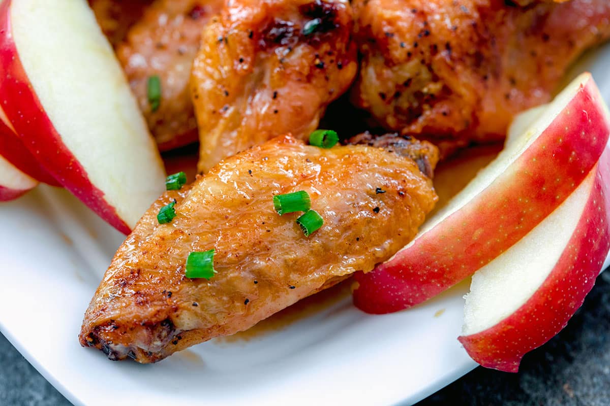 Landscape head-on view of apple cider chicken wings on platter with sliced apples and chives.