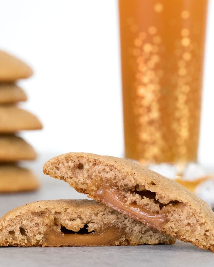 Head-on closeup view of an apple cider cookie cut in half with caramel oozing out with a stack of more cookies and glass of apple cider in the background