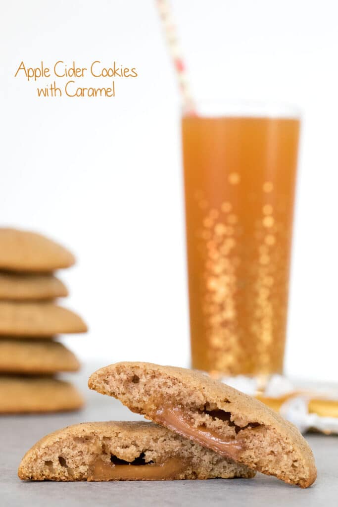 Head-on view of an apple cider cookie cut in half with caramel oozing out with a stack of more cookies and glass of apple cider in the background and recipe title at top