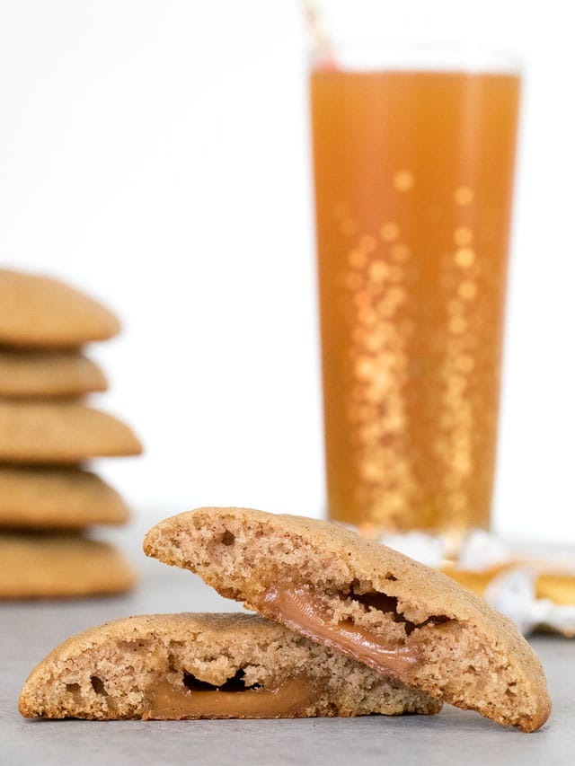 Head-on view of an apple cider cookie cut in half with caramel oozing out with a stack of more cookies and glass of apple cider in the background