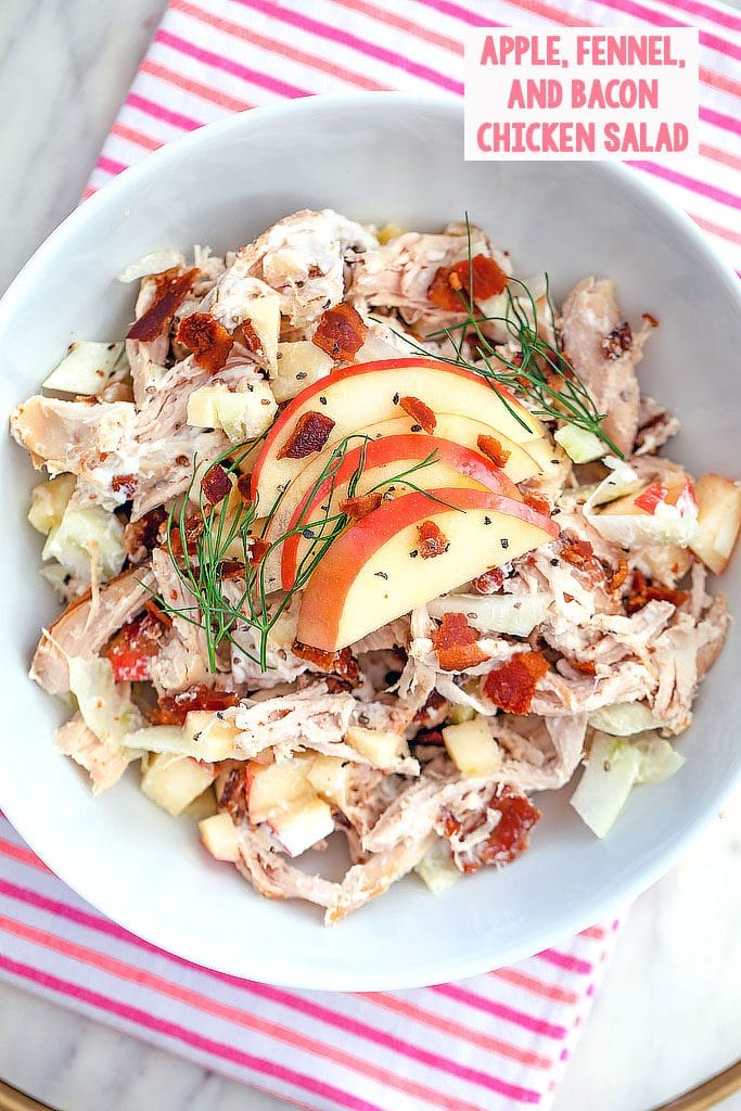 Overhead view of apple, fennel, and bacon chicken salad topped with sliced apples and fennel fronds on a pink and red striped towel with recipe title at the top