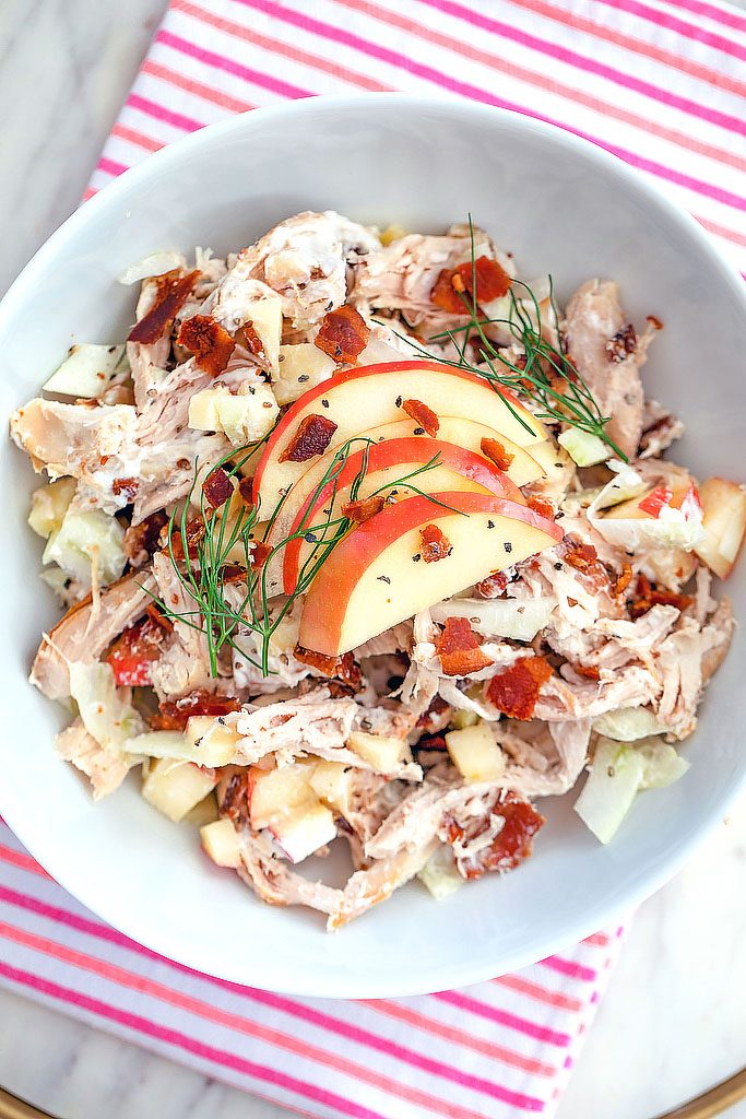 Overhead view of apple, fennel, and bacon chicken salad topped with sliced apples and fennel fronds on a pink and red striped towel