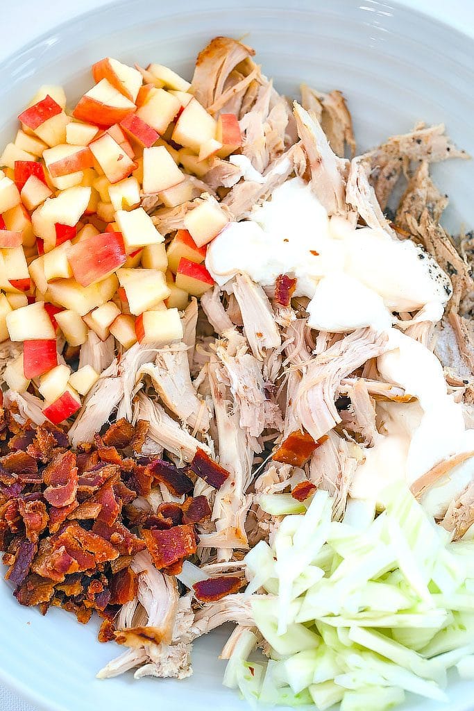 Overhead view of a white bowl containing shredded chicken, diced apple, crumbled bacon, sliced fennel, greek yogurt, and mayo