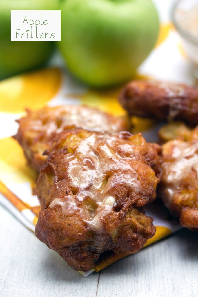 Head-on view of an apple fritter with icing with more fritters and granny smith apples in the background and recipe title at top