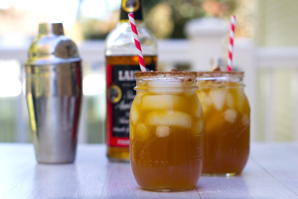 Landscape head-on view of two apple ginger margaritas in mason jars with cinnamon rims and red and white striped straws with bottle of apple brandy and cocktail shaker in the background