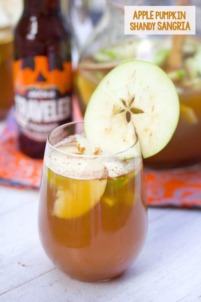 Head-on view of a glass of apple pumpkin shandy sangria with an apple ring garnish and a bottle of pumpkin shandy and pitcher of the cocktail in the background with the recipe title at the top