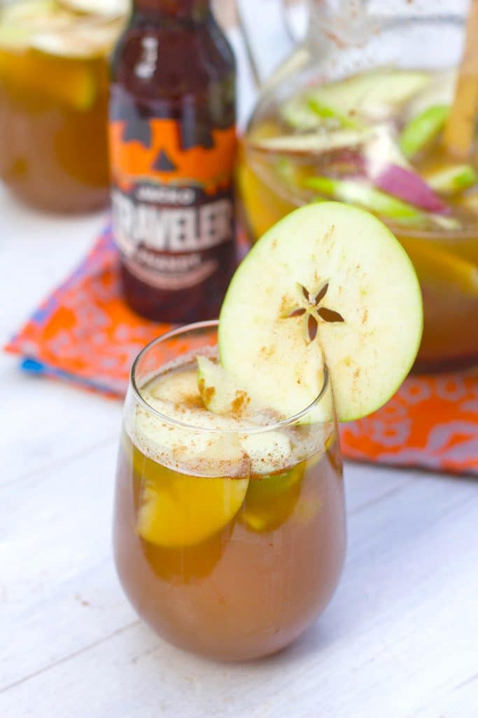 Glass of apple pumpkin shandy sangria with an apple garnish and a bottle of pumpkin shandy and pitcher of the cocktail in the background