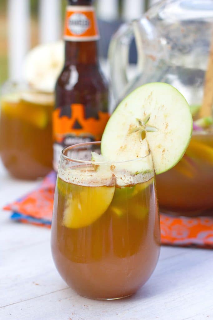 Head-on view of a glass of apple pumpkin shandy sangria with an apple ring garnish and a bottle of pumpkin shandy and pitcher of the cocktail in the background