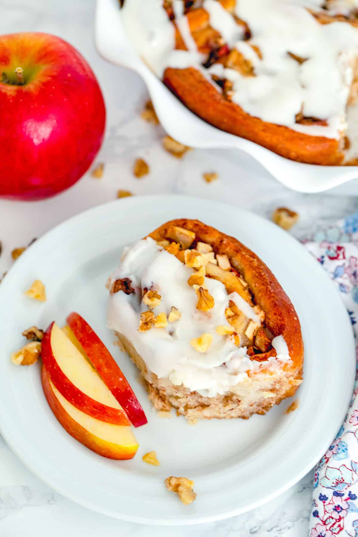 Overhead view of an apple walnut cinnamon roll on a plate with sliced apples with whole apple and dish of cinnamon rolls in the background with walnuts scattered all around.