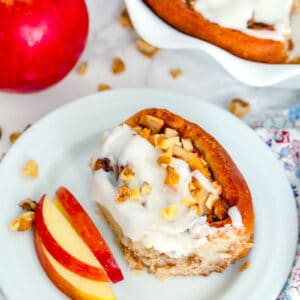 Overhead view of an apple walnut cinnamon roll on a plate with sliced apples with full platter of cinnamon rolls, apple, and walnuts scattered around
