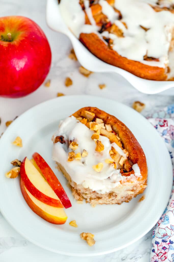 Overhead view of an apple walnut cinnamon roll on a plate with sliced apples with whole apple and dish of cinnamon rolls in the background with walnuts scattered all around