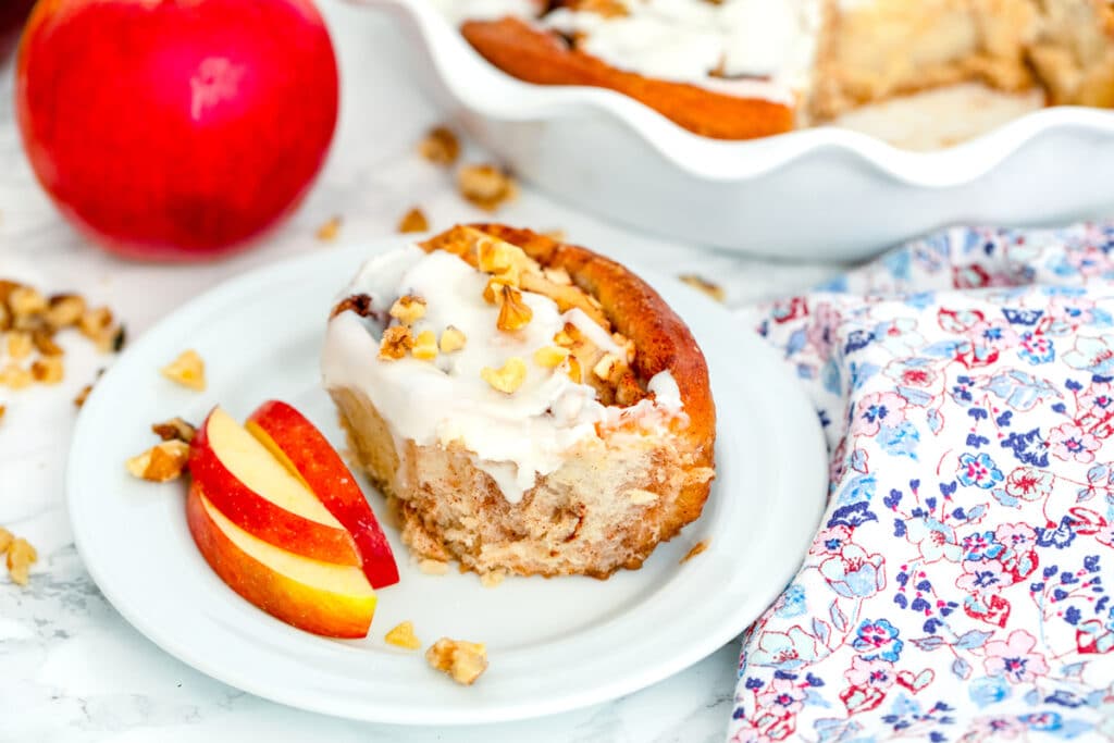 Landscape head-on view of an apple walnut cinnamon roll on a small plate with sliced apple with apple and dish of more cinnamon rolls in the background with walnuts all around