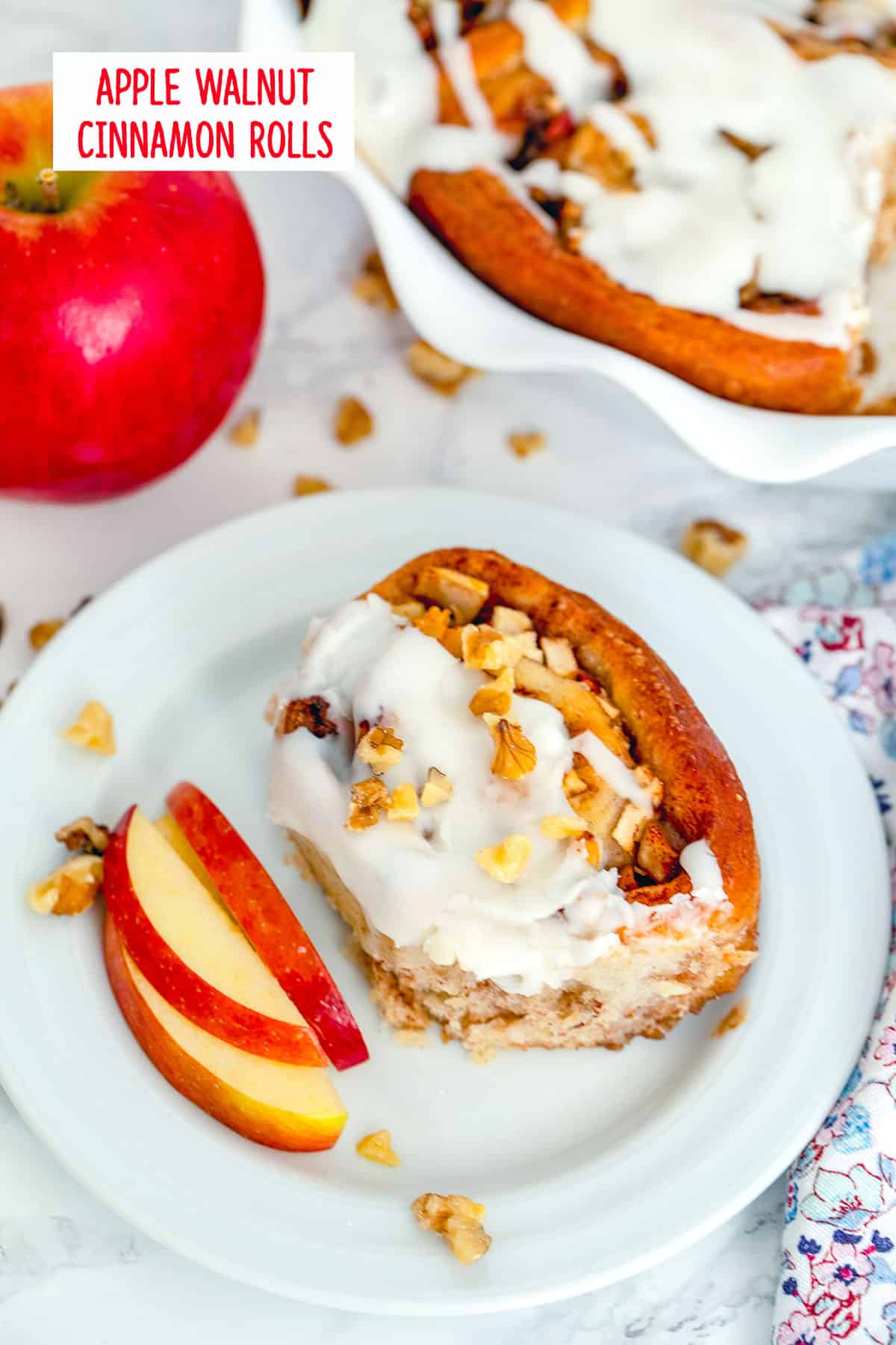 Overhead view of an apple walnut cinnamon roll on a plate with sliced apples with whole apple and dish of cinnamon rolls in the background with walnuts scattered all around and recipe title at top.