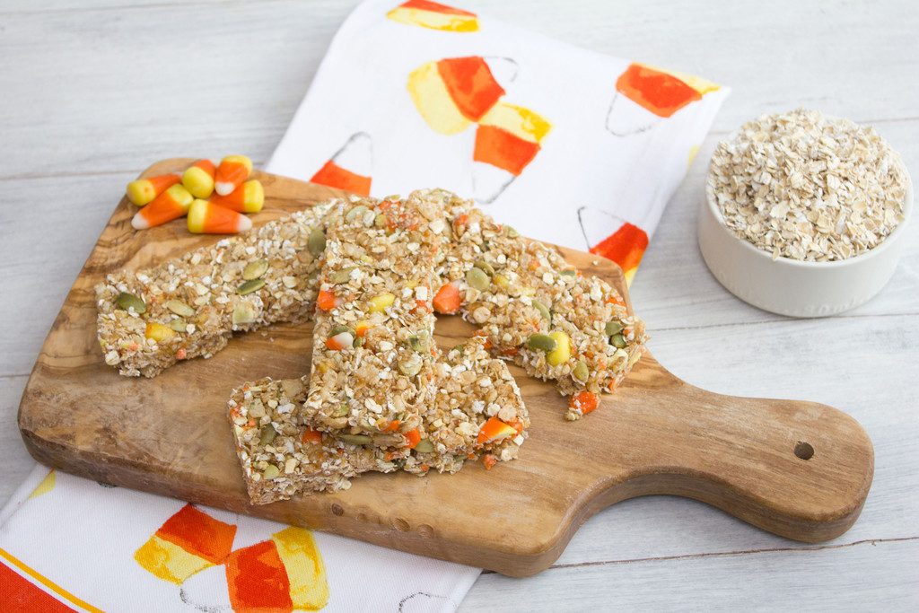 Overhead landscape view of wooden board with candy corn granola bars on it with some candy corn on a candy corn tea towel with a cup of oats in the background