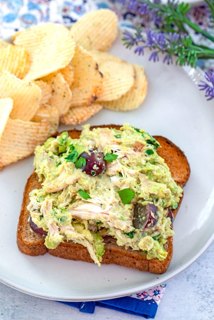 Overhead view of a slice of bread topped with avocado chicken salad with grapes on a plate with chips on the side