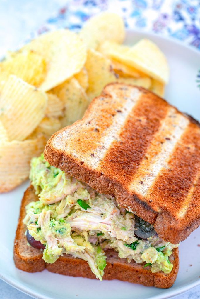 Overhead view of avocado chicken salad sandwich with top slice of break slightly removed to show chicken salad filling on a plate with chips on the side