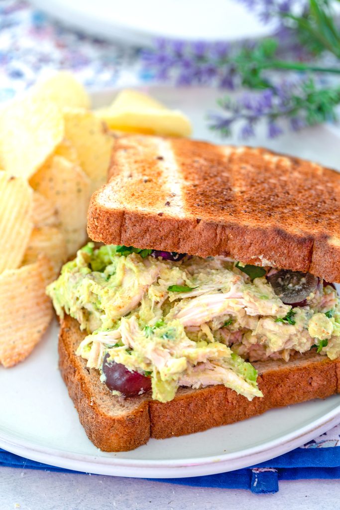 Head-on view of avocado chicken salad sandwich with top slice of bread slightly removed to show chicken salad on a plate with chips on the sid