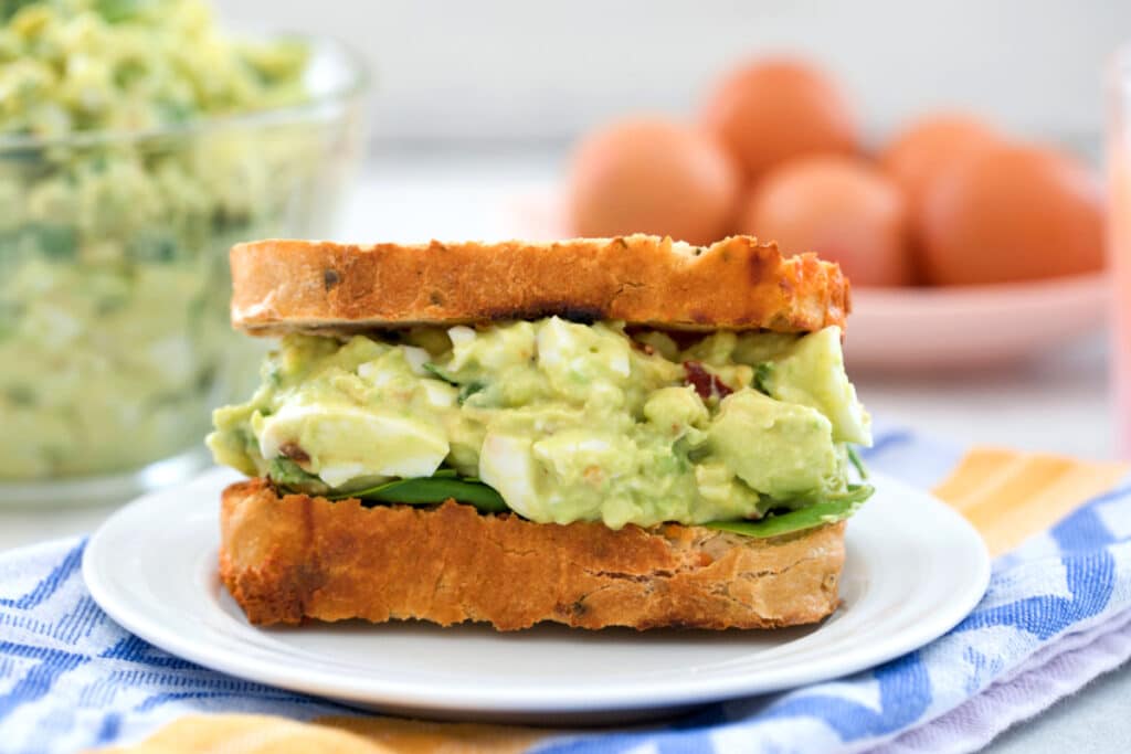 Landscape head-on view of a sandwich filled with avocado egg salad with bowl of egg salad and brown eggs in background