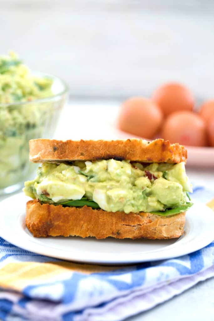 Head-on closeup view of an avocado egg salad sandwich with spinach with bowl of egg salad and eggs in the background