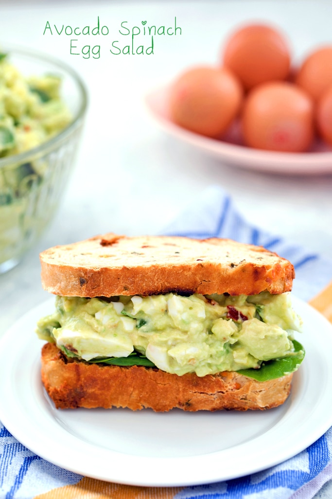 Head-on view of a sandwich packed with avocado egg salad with spinach with bowl of avocado egg salad and eggs in the background and recipe title at top