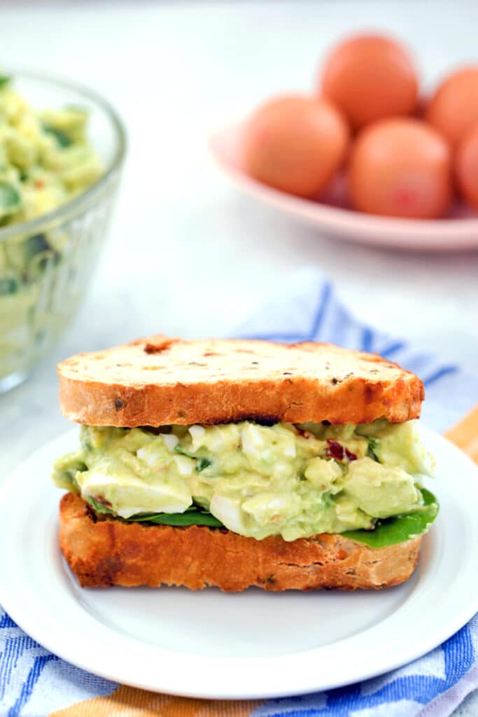 Head-on view of a sandwich packed with avocado egg salad with spinach with bowl of avocado egg salad and eggs in the background