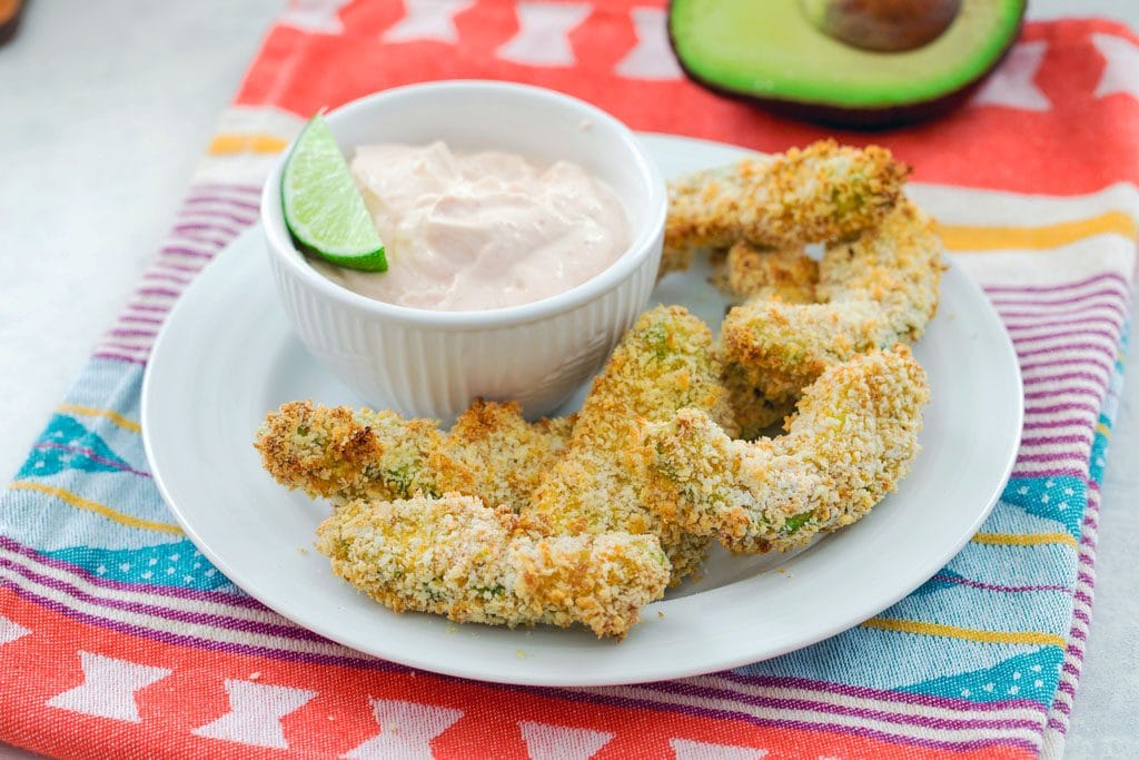 Horizontal shot from a distance of panko-coated avocado fries with a white bowl of sriracha dipping sauce with a lime in it and half an avocado in the background