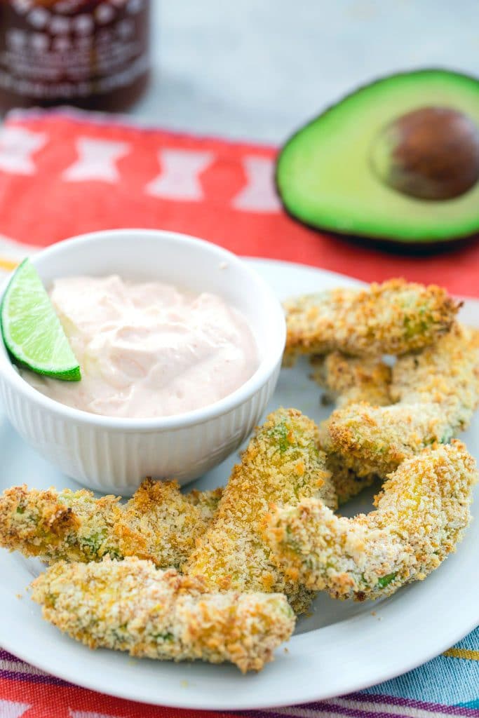 Overhead shot of panko-coated avocado fries with a bowl of sriracha dipping sauce with a lime in it and half an avocado and a sriracha bottle in the background 