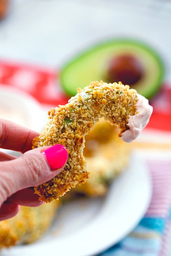 Close up of a hand with pink nail polish holding a panko-coated avocado slice with some sriracha dipping sauce on the top of it