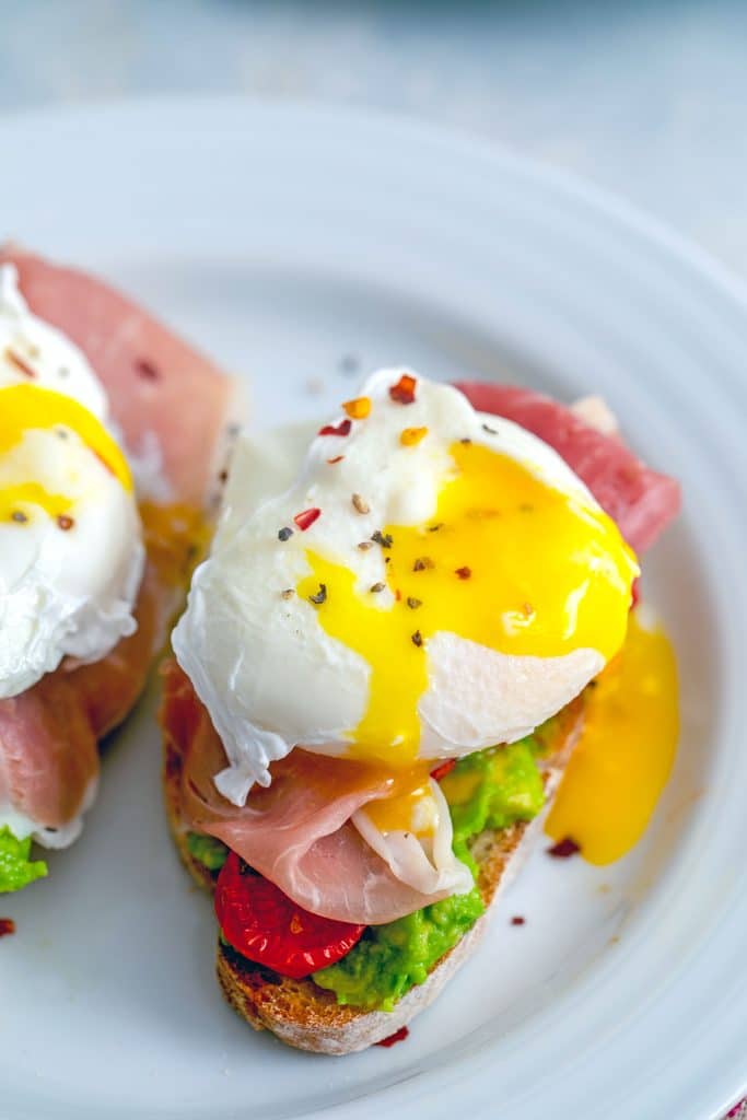 Overhead view of avocado egg toast with tomatoes and prosciutto and yolk running out of egg onto white plate