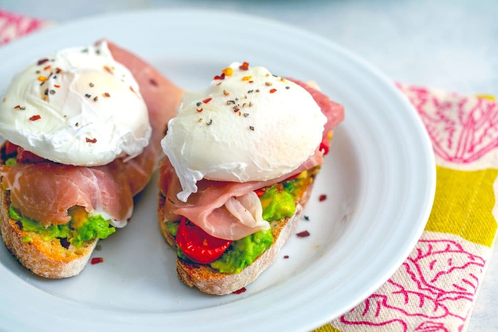 Landscape view of two slices of avocado egg toast on a white plate with tomatoes and prosciutto with eggs not yet broken