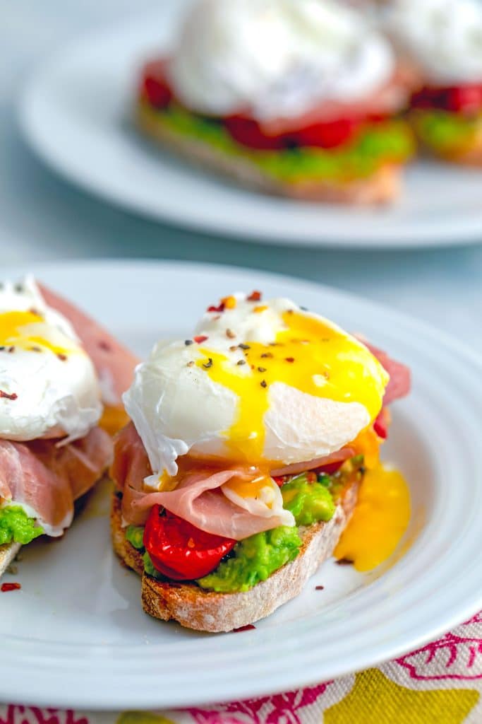 Head-on view of slice of avocado egg toast with runny egg, roasted tomatoes, and avocado with another plate in the background