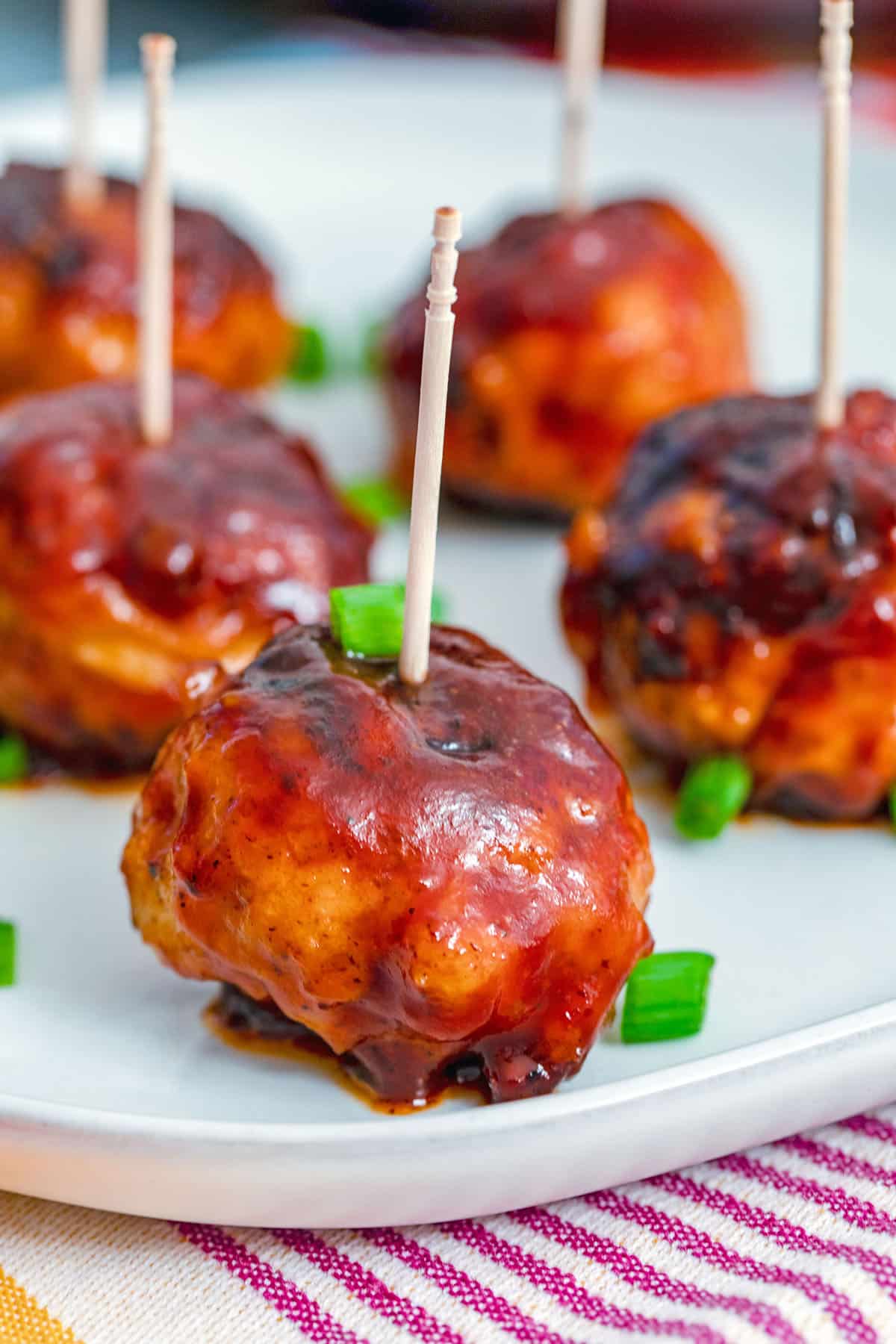 Head-on closeup view of a BBQ Chicken meatball with a toothpick in it with more meatballs in the background and chopped scallions all around.