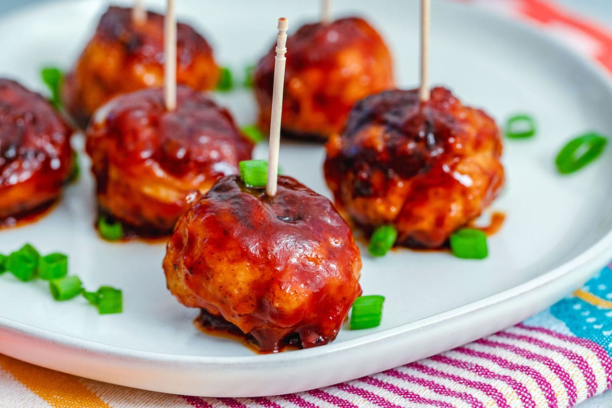 Landscape head-on view of multiple BBQ chicken meatballs with toothpicks on plate with chopped scallions all around.