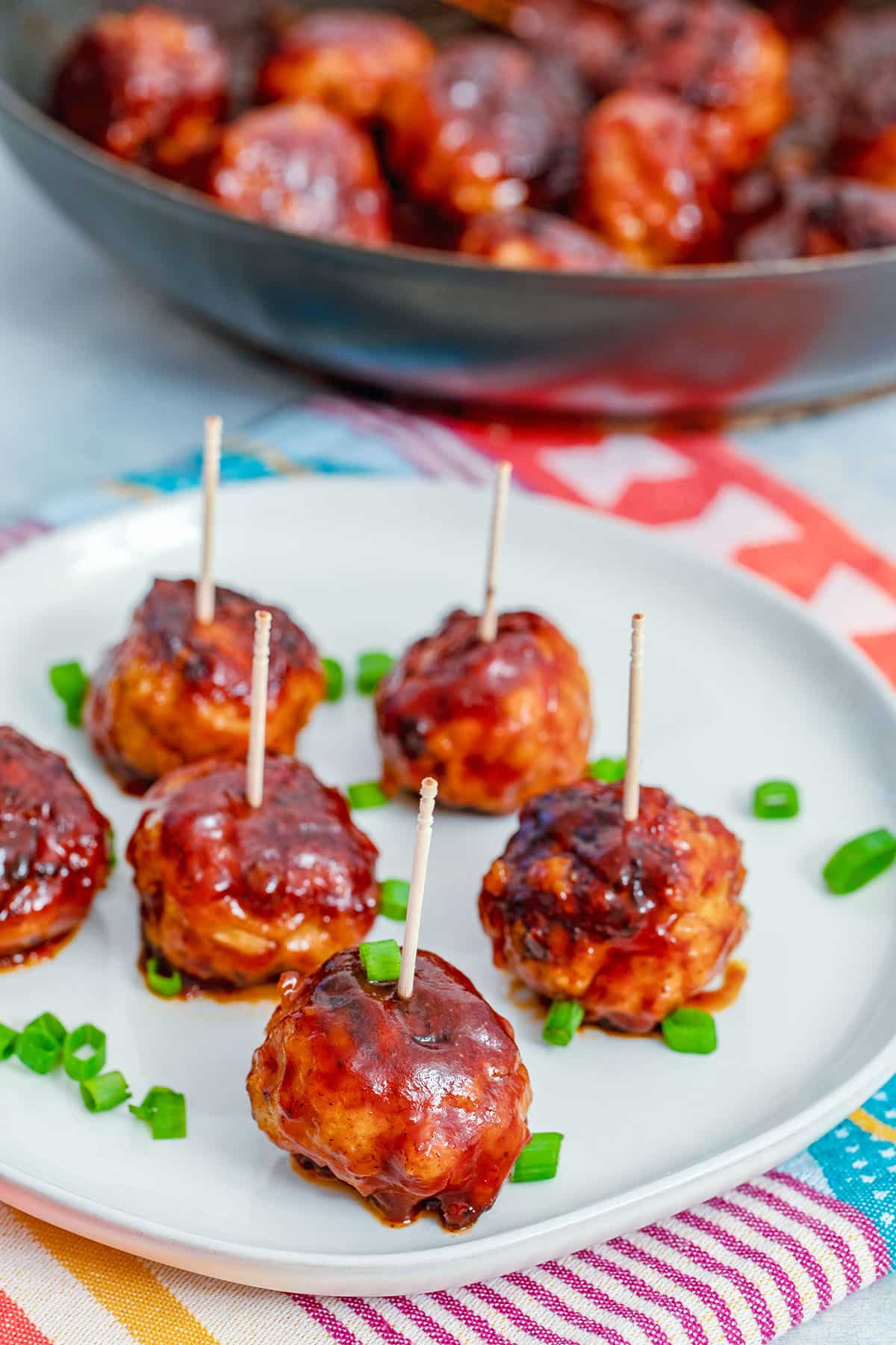 View of a plate filled with BBQ chicken meatballs with toothpicks and chopped scallions with skillet with more BBQ chicken meatballs in the background.