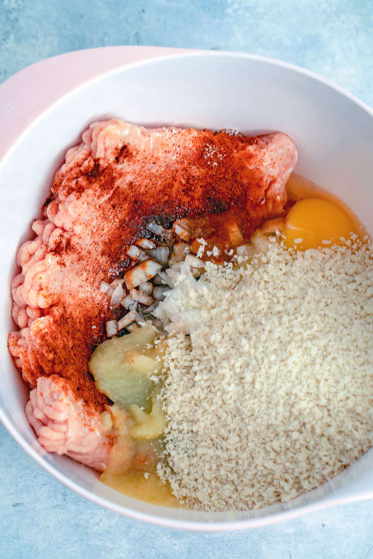 Overhead view of ingredients in mixing bowl, including ground chicken, panko, egg, onion, applesauce, and spices.