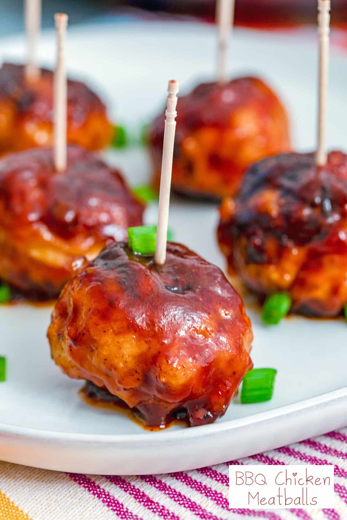 Head-on closeup view of a BBQ Chicken meatball with a toothpick in it with more meatballs in the background, chopped scallions all around, and recipe title at bottom.