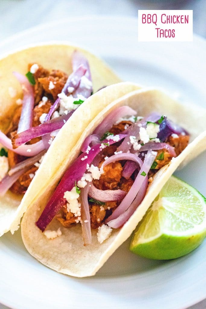 Head-on view of two BBQ chicken tacos topped with red onions on a white with a lime wedge and recipe title at top