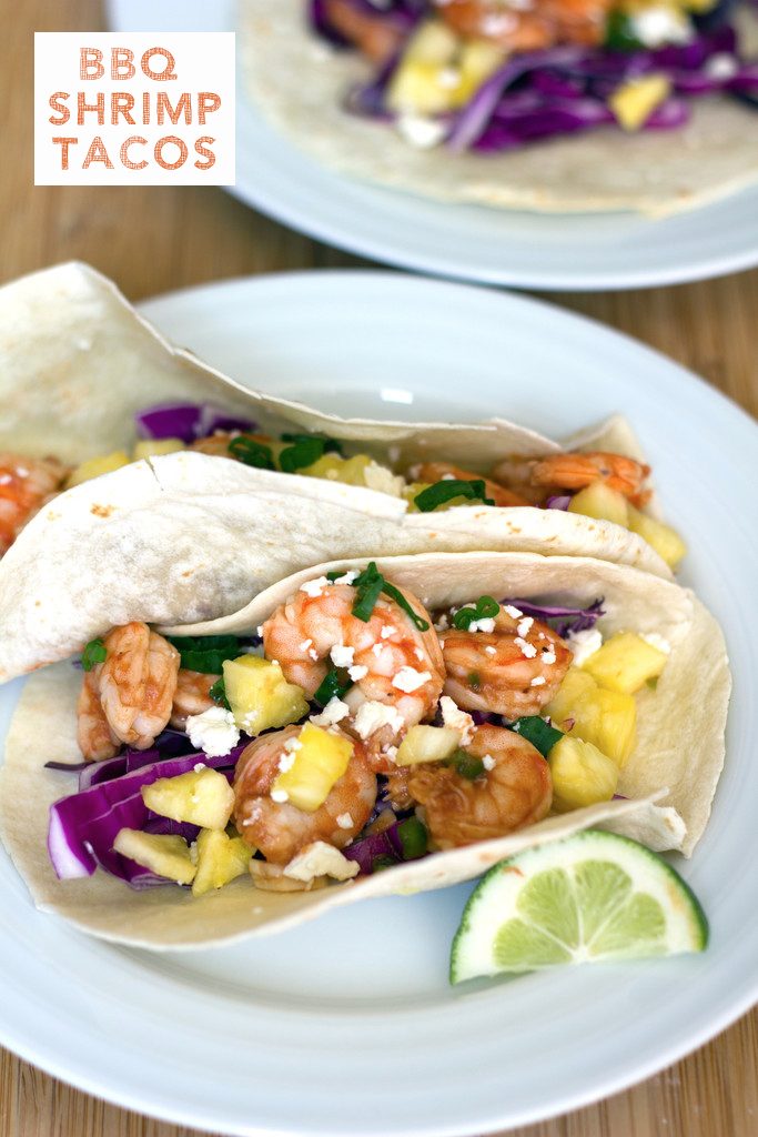 Head-on view of two BBQ shrimp tacos on a white plate with a lime wedge with second plate in the background and recipe title at top