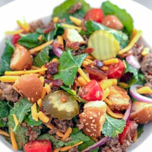 Overhead close-up view of bacon cheeseburger kale salad including baby kale, ground beef, shredded cheddar, dill pickle rounds, chopped tomatoes, red onion, crumbled bacon, and hamburger bun croutons in a white bowl with recipe title at top